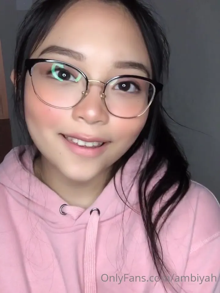 Mae in a pink hoodie and glasses, licking her lips and flashing her tongue