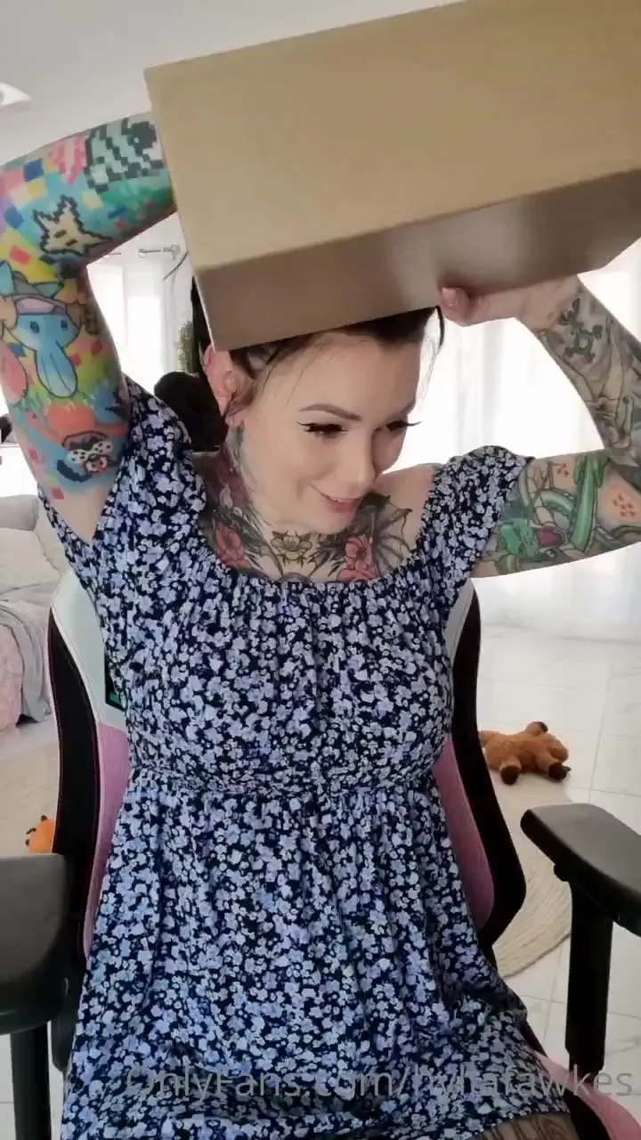 Hylia tears into a cardboard package in her navy floral dress, ink flexing as she reads the label