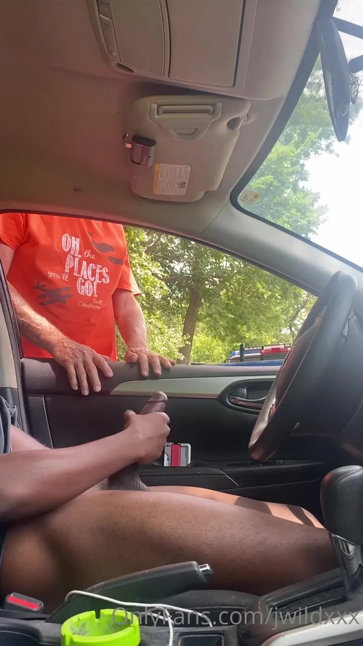 Jwildxxx in an open car door, jerking off hungry while a grey-haired stranger in an orange tee takes his thick dick in hand