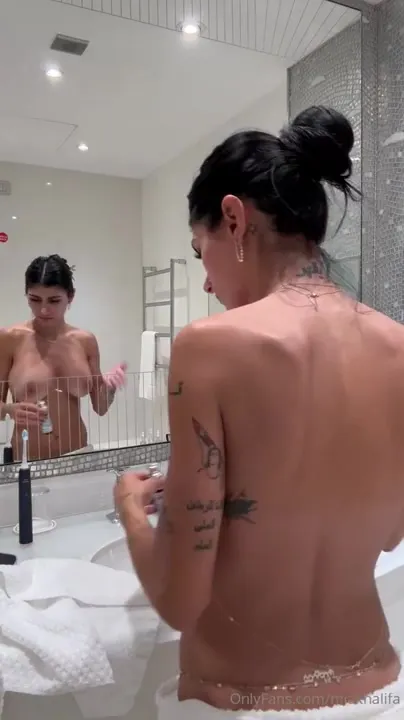 In a low white towel, Mia Khalifa brushes her teeth and lets her orange robe fall open