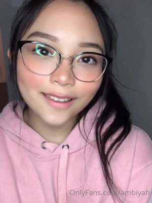 Mae in a pink hoodie and glasses, licking her lips and flashing her tongue